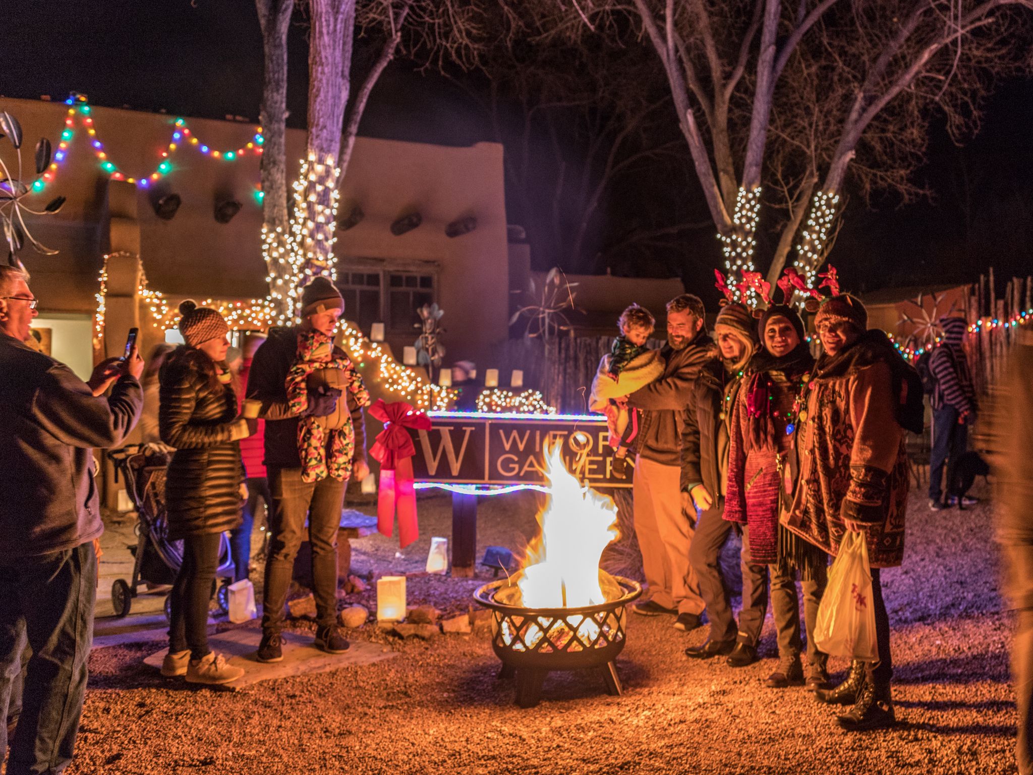 http://Group of people bundled in warm clothing enjoying a lively winter gathering at night, with a fire pit, festive lights in front of a shopfront.