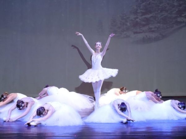 A scene from a ballet performance featuring a central dancer in a white tutu standing with arms gracefully positioned, surrounded by dancers lying on the stage in white costumes, depicting a serene and artistic tableau.