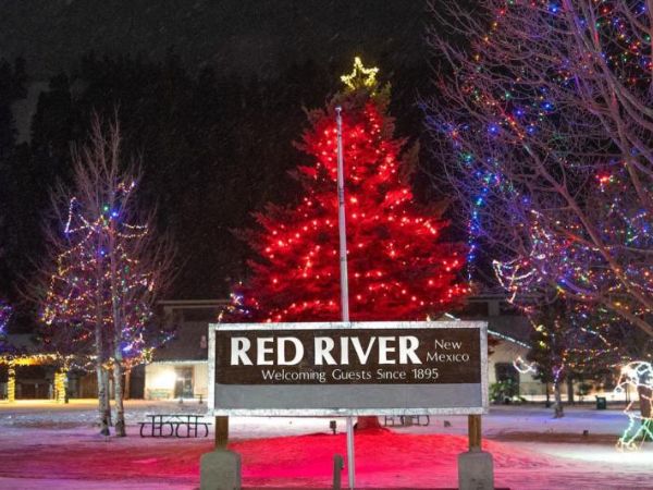 A vibrant night scene in Red River, New Mexico, showcasing a brightly decorated Christmas tree with a star on top and illuminated trees along a park.