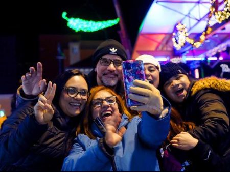 A group of people standing next to a stage, one is holding a phone, illuminated by festive lights.