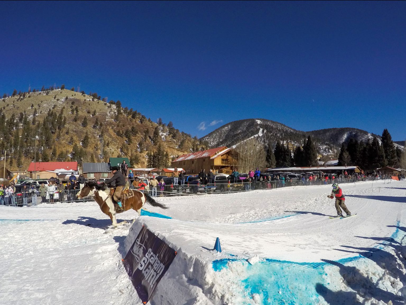 Scene at a winter sports event with a person skiing down a snowy street being pulled by another person riding a horse. Spectators line the course, with mountainous terrain in the background under a clear blue sky.