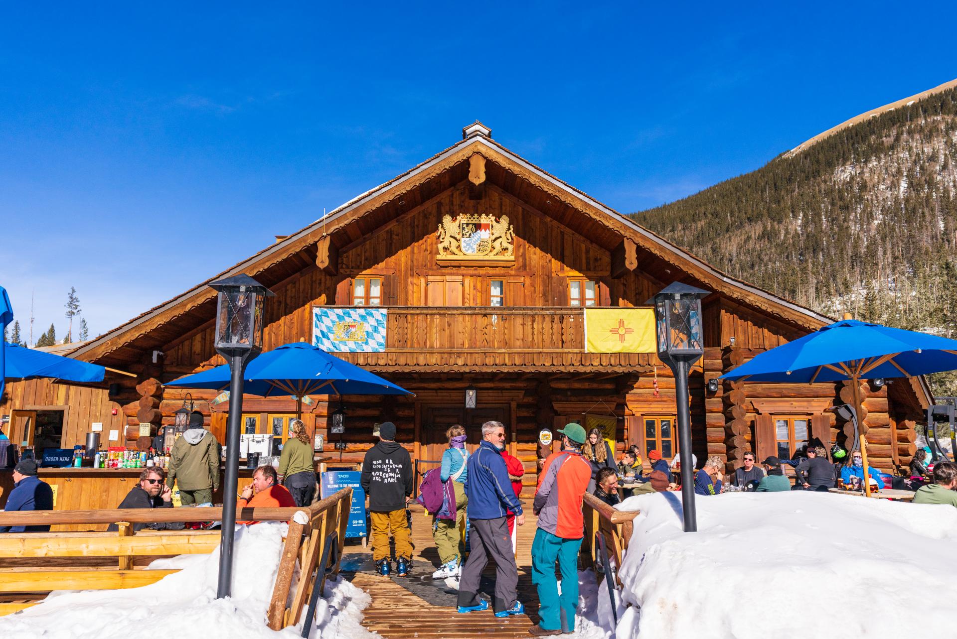 People gather outside a wooden ski lodge with blue umbrellas, under a clear blue sky in a snowy mountain setting.