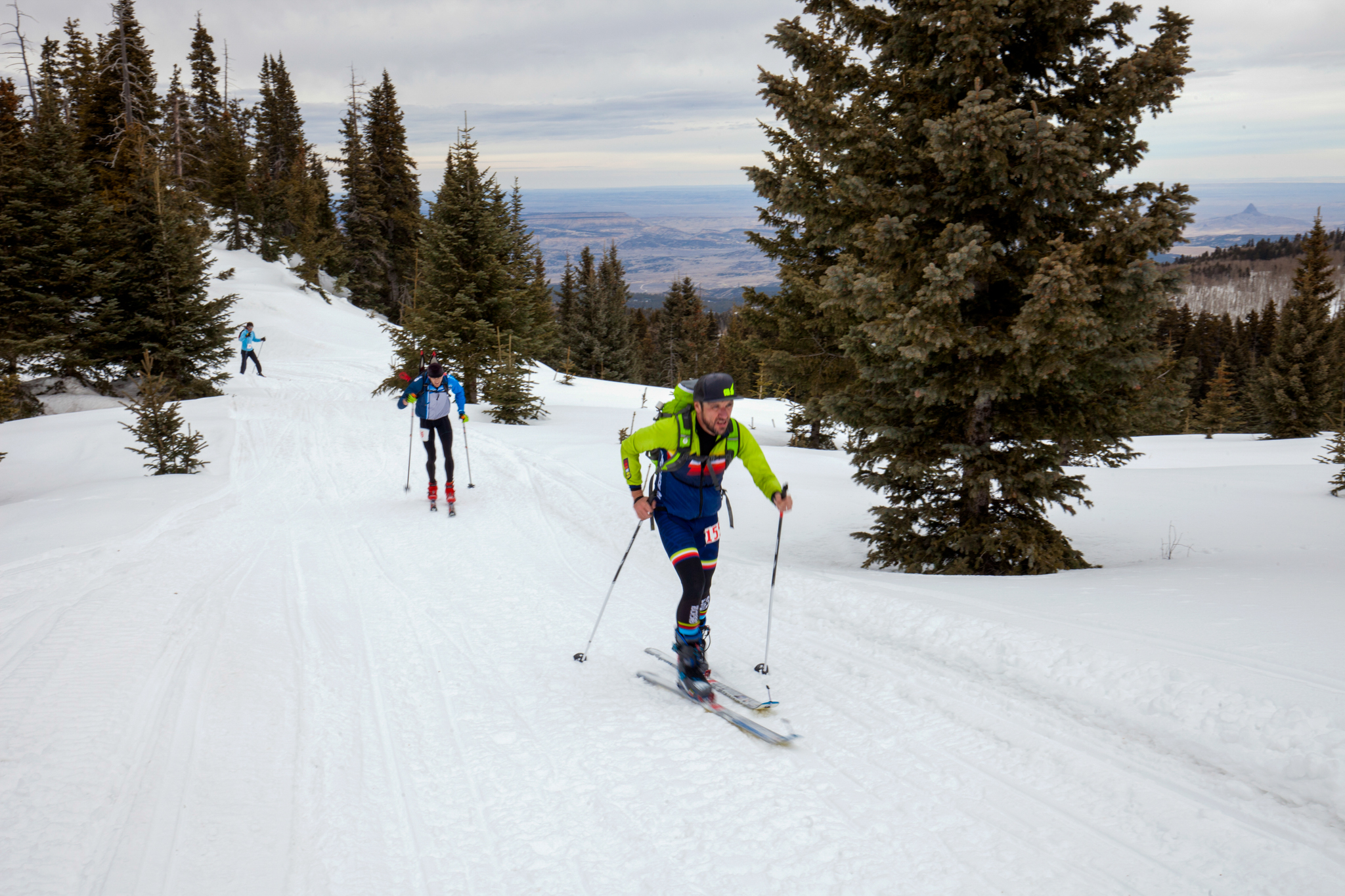 People cross-country skiing along a tree-lined trail with a scenic snow-covered landscape in the background.