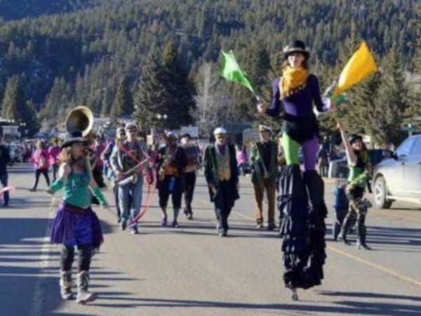 A group of people participating in a street parade, some playing musical instruments and others waving colorful flags, with mountains in the background and onlookers lining the streets.