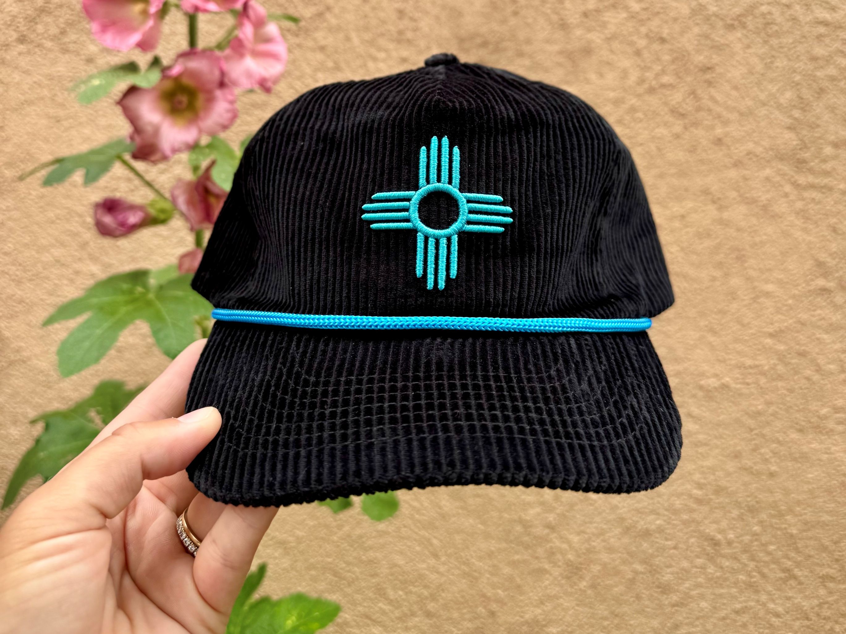 A hand holding a black corduroy hat with a Zia symbol embroidered in turquoise, positioned in front of a wall with a hollyhock flower beside it.