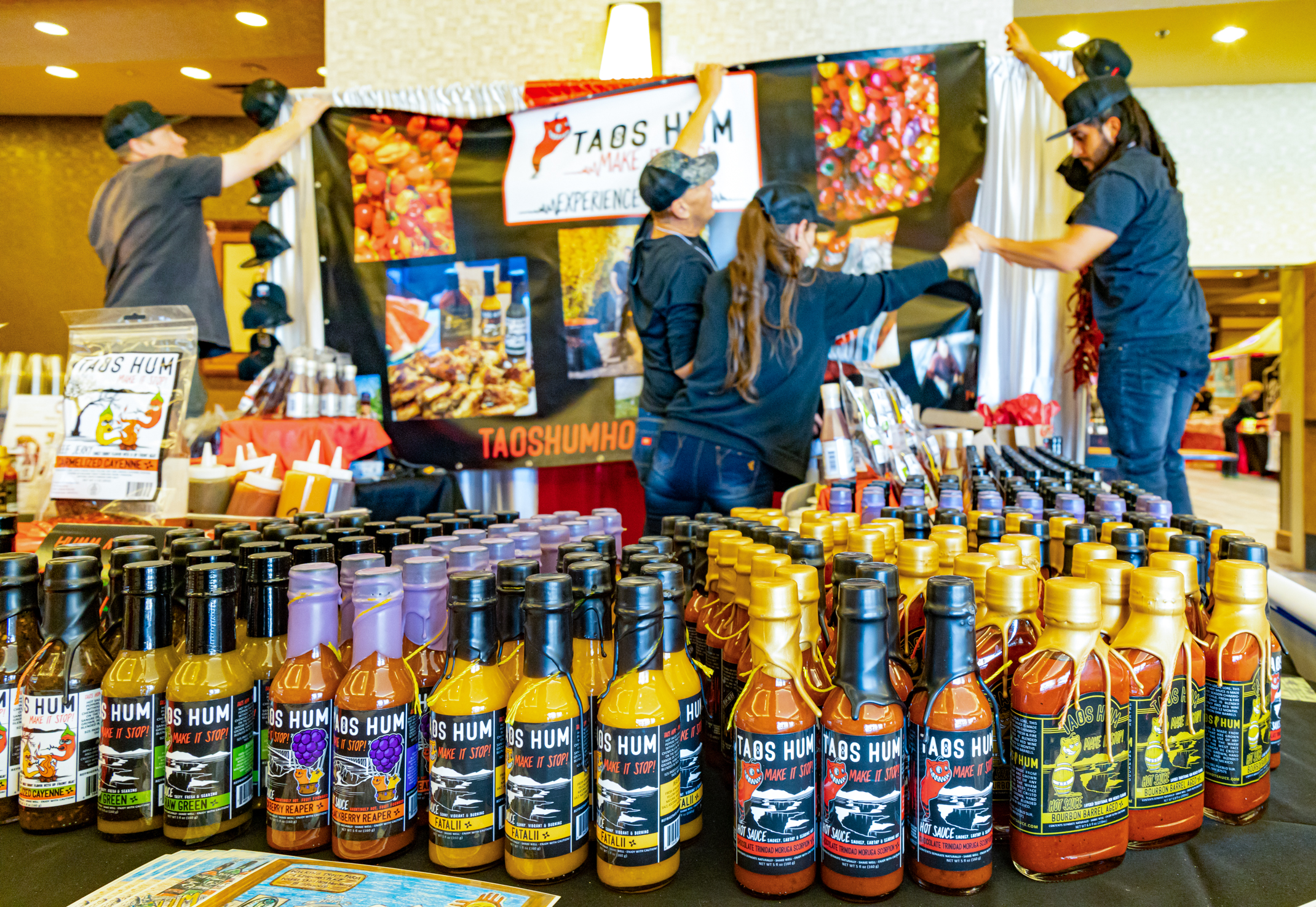 Bottles of colorful sauces sit on a table as people in the background hang a large banner with product names in the background
