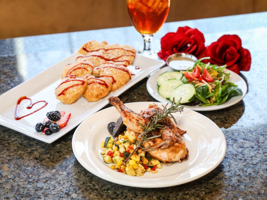 A dining table setting with three dishes: grilled chicken on a bed of mixed vegetables garnished with rosemary, a plate of croissants drizzled with sauce accompanied by berries, and a fresh salad with cucumber and tomato slices. Red roses and an iced tea are in the background.