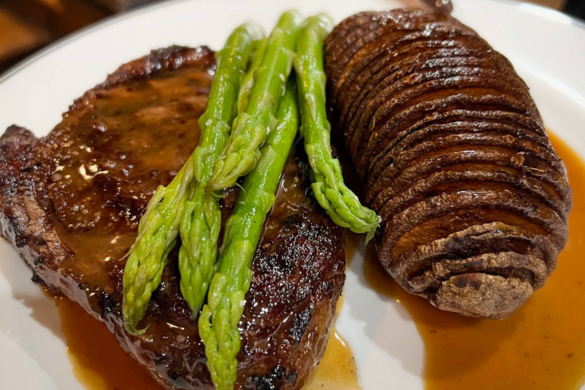 A plate featuring a grilled steak topped with asparagus spears, accompanied by a seasoned and cut potato, drizzled with a brown sauce.