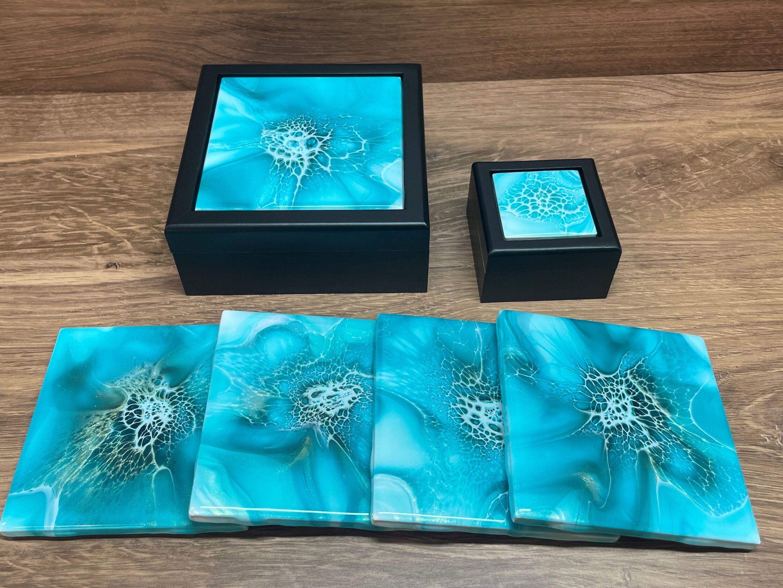 A collection of items featuring a vibrant turquoise design with white abstract patterns. There are two black boxes, one larger and one smaller, and four rectangular coasters displayed in front, all on a wooden surface.