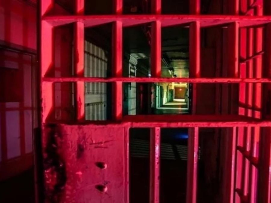 A dimly lit prison corridor with cell bars in the foreground, illuminated by red lighting. The hallway extends into darkness, creating a dramatic perspective.