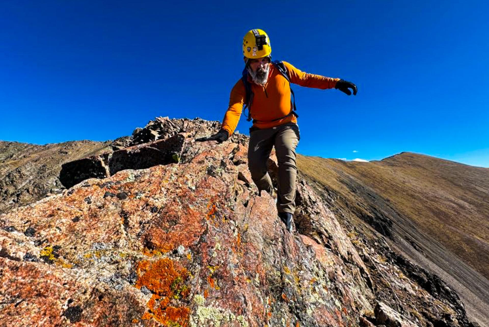A person wearing an orange top and yellow helmet is climbing on a rocky mountain ridge under a clear blue sky.