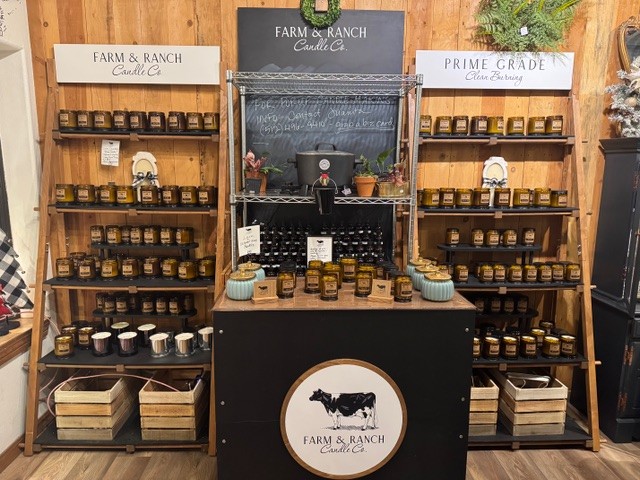 A display of "Farm & Ranch Candle Co." products is showcased on shelves with jars of candles. The setup includes a chalkboard with writing and decorative plants.