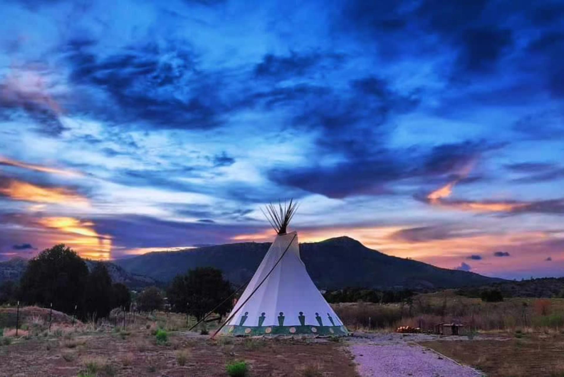 A tipi stands in an open field at sunset, with a vivid blue and orange sky overhead. Rolling hills and scattered trees form the background.