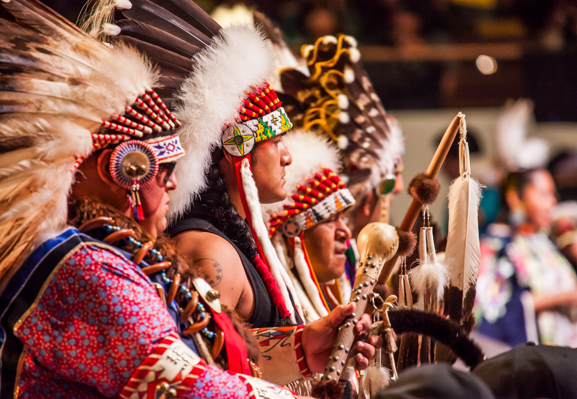 A group of people in traditional Indigenous attire stand in a line playing instruments.