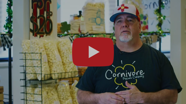 A man sitting in front of a rack filled with bags of popcorn with various signs in the background.