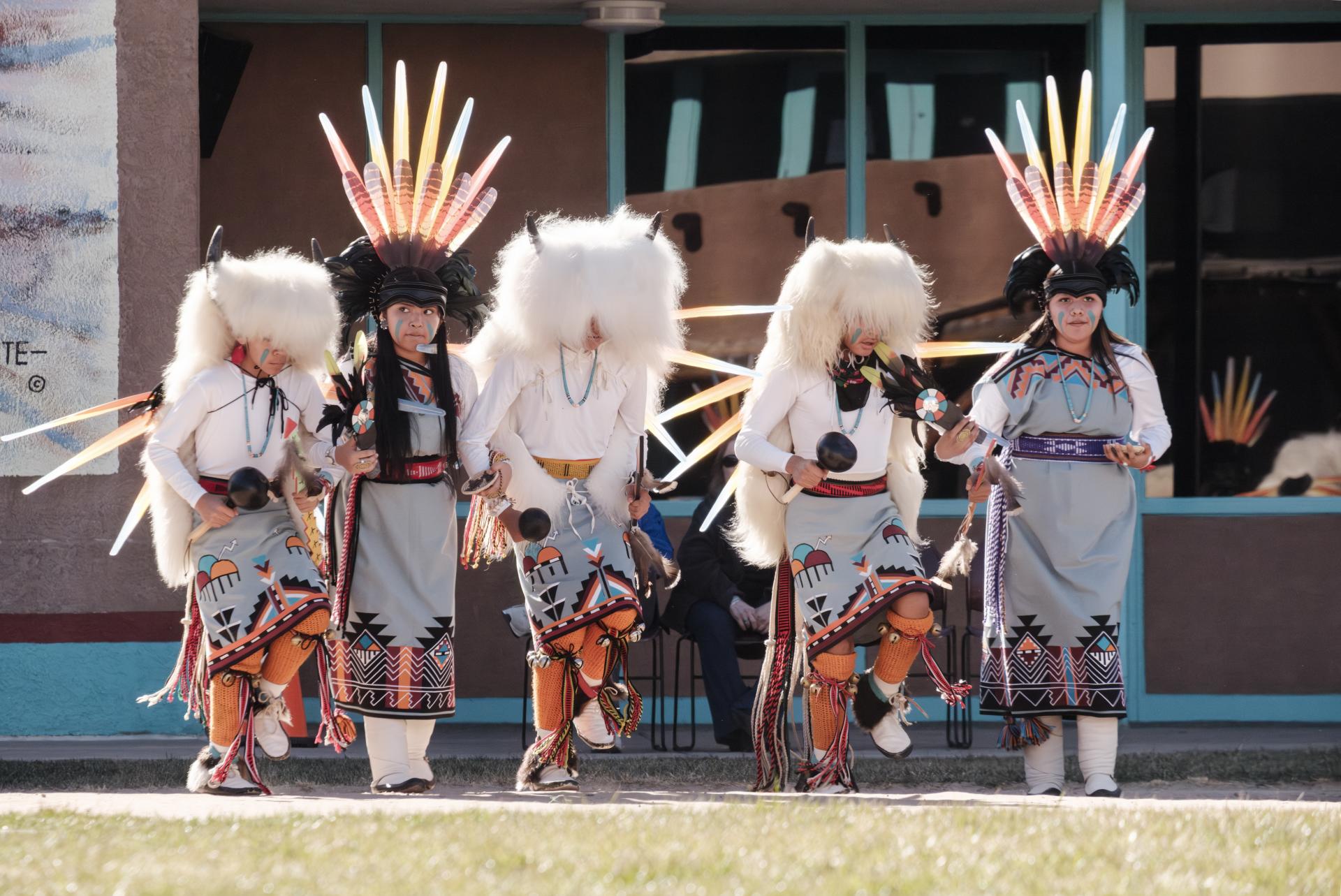 A group of Indigenous Peoples are performing a traditional dance outside a building, wearing ornate regalia with colorful feathered headdresses and decorated garments.