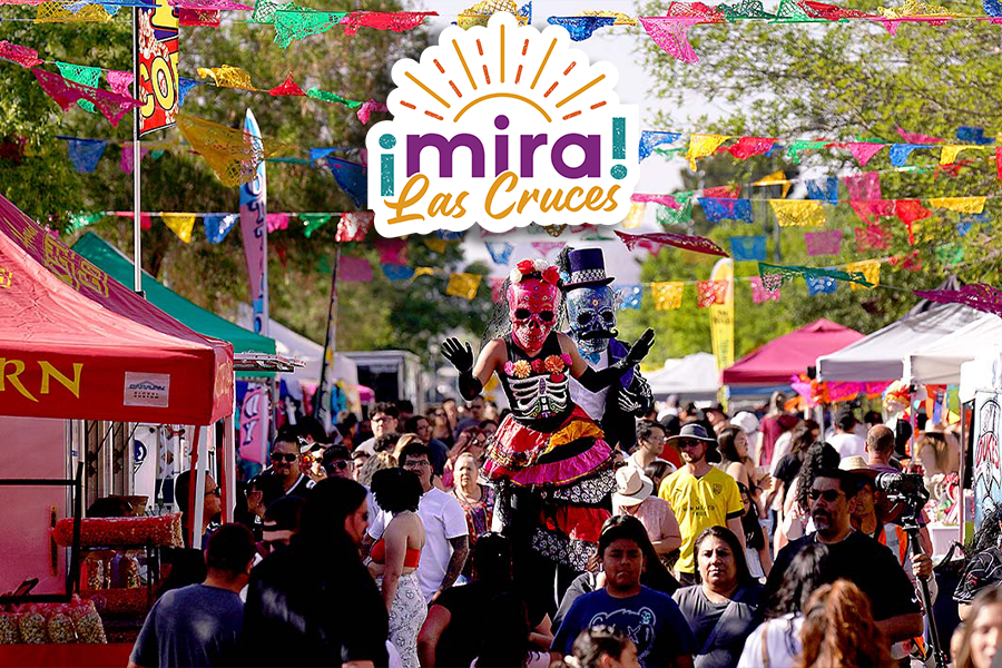 A lively festival scene with colorful papel picado banners hanging overhead. In the center, two performers in vibrant, traditional Dia de los Muertos costumes stand out.  A prominent sign at the top reads "¡Mira! Las Cruces."