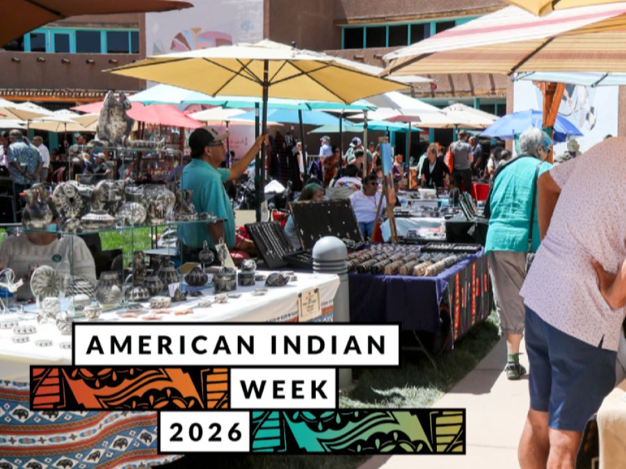A bustling outdoor market under colorful umbrellas with numerous tables displaying various crafts and jewelry. A banner in the foreground reads "American Indian Week 2026."
