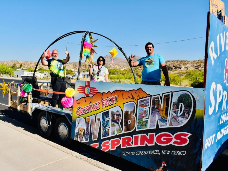 A parade float decorated with a colorful "Greetings from Riverbend Hot Springs" sign, featuring vibrant desert imagery and the text "Truth or Consequences, New Mexico." Three individuals stand on the float, surrounded by vivid paper lanterns and festive decorations.