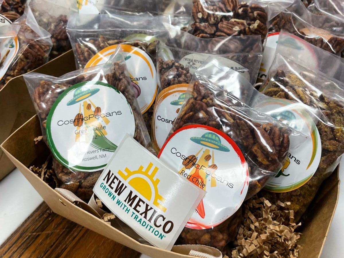 A box filled with various packages of "Cosmic Pecans," each labeled with a colorful sticker. At the front, a sign reads "New Mexico, Grow With Tradition." The packages contain pecans displayed in clear bags.