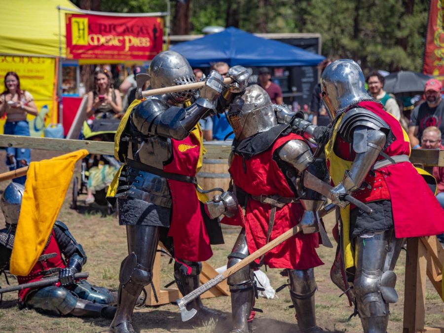 Three individuals in medieval armor are engaging in a mock battle at a Renaissance fair, while a crowd watches. A participant is seated on the ground. 