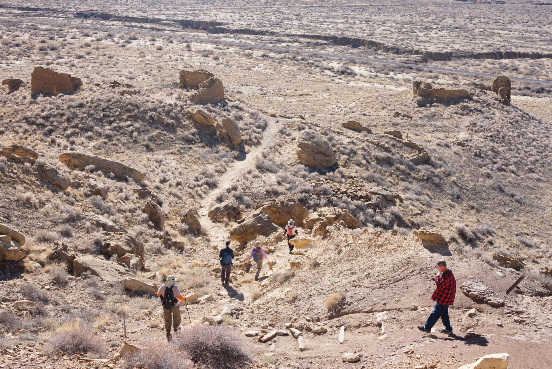 A group of people hiking through a desert landscape with scattered ruins at Chaco Canyon, New Mexico. The terrain is rocky and arid with sparse vegetation.