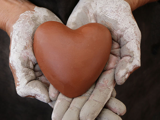 Hands covered in clay gently holding a smooth, heart-shaped object made of clay.