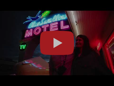 Two people beneath a large neon sign that reads Blue Swallow Motel, the people are bathed in warm light.