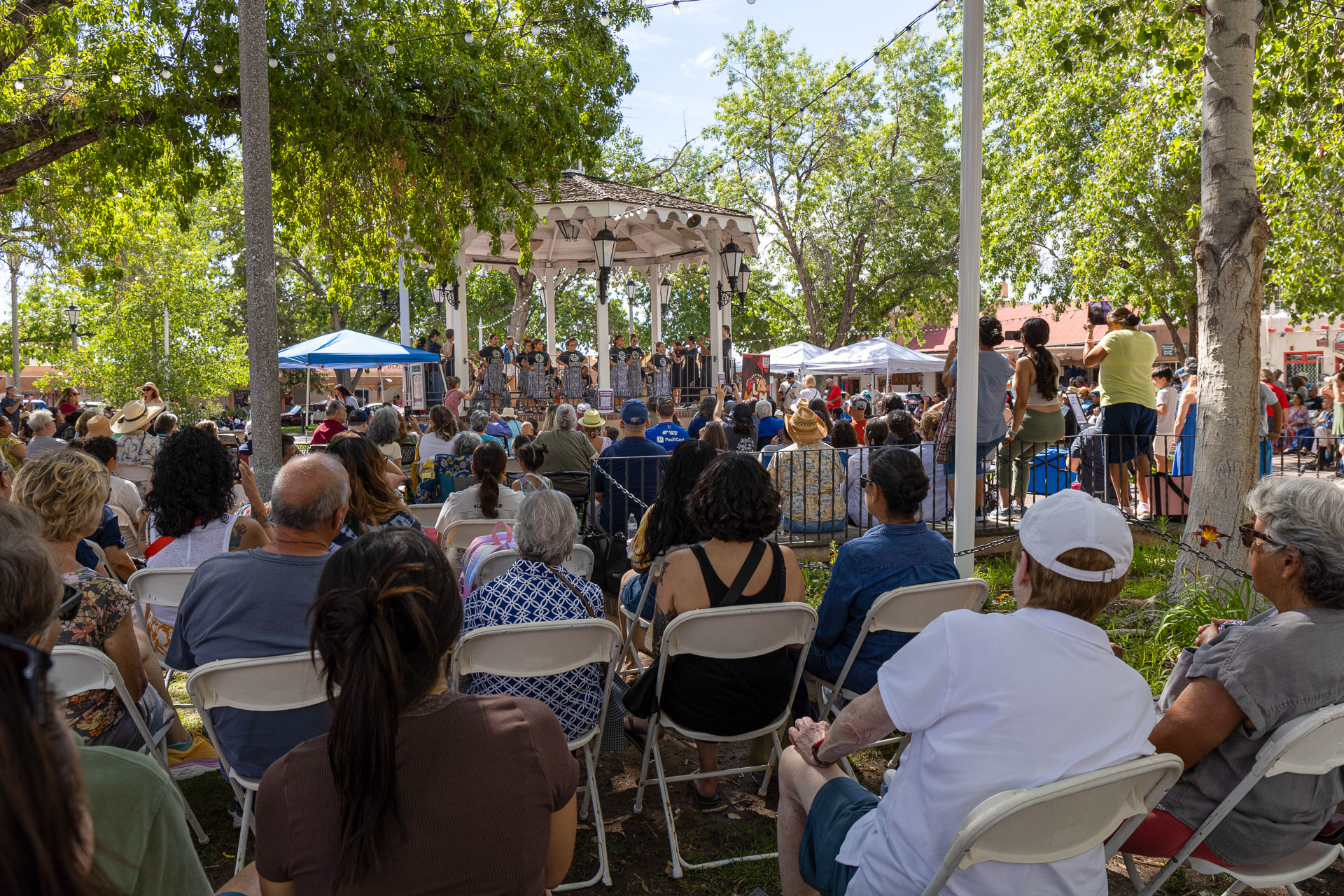A diverse group of people is seated outdoors, facing a gazebo stage where a performance is taking place. The setting is surrounded by trees, creating a shaded area. Some attendees are standing, and tents are visible in the background.