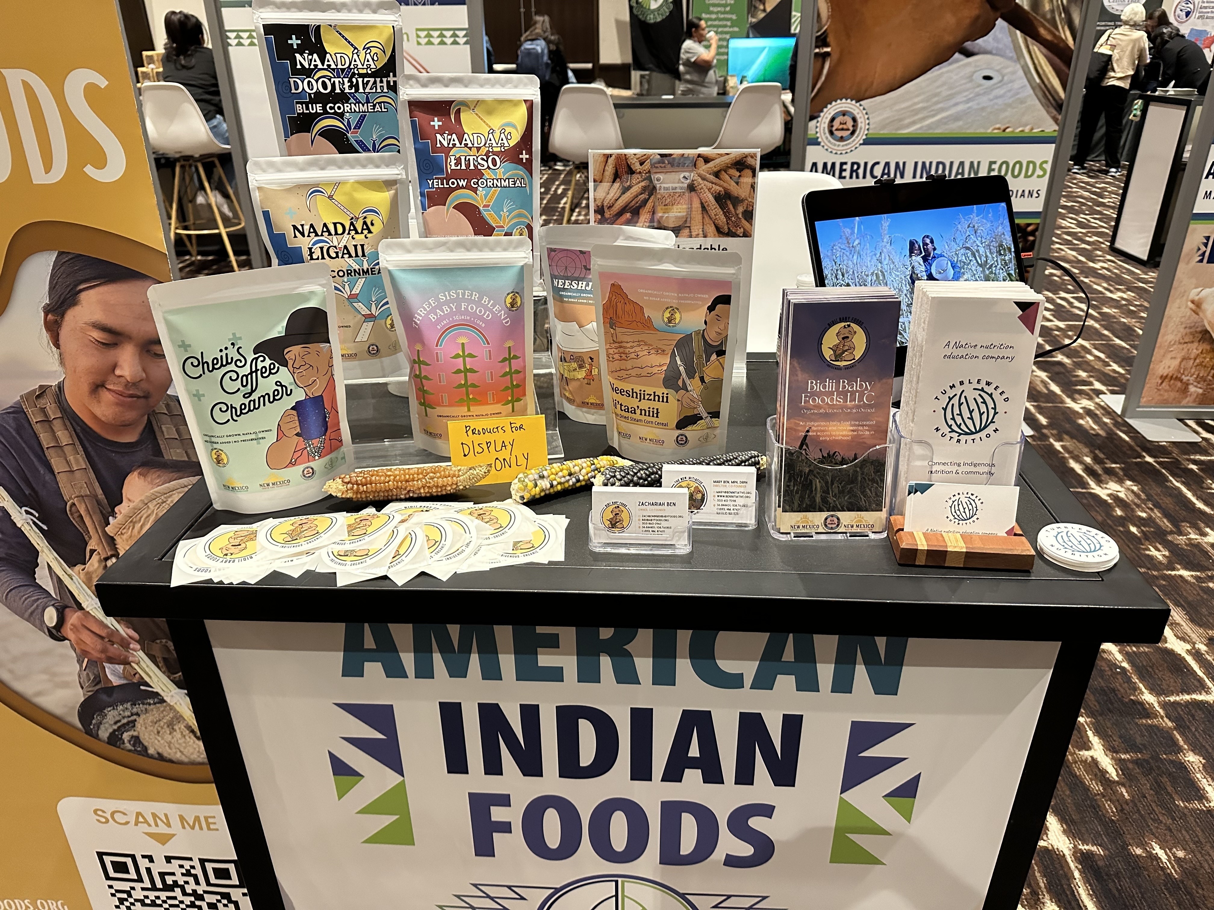 Exhibit booth displaying various American Indian foods, including Sunflower Foods and Tanka. There are packages of popcorn, corn flour, and bags with colorful labels. A banner reads "American Indian Foods," and QR codes are present.