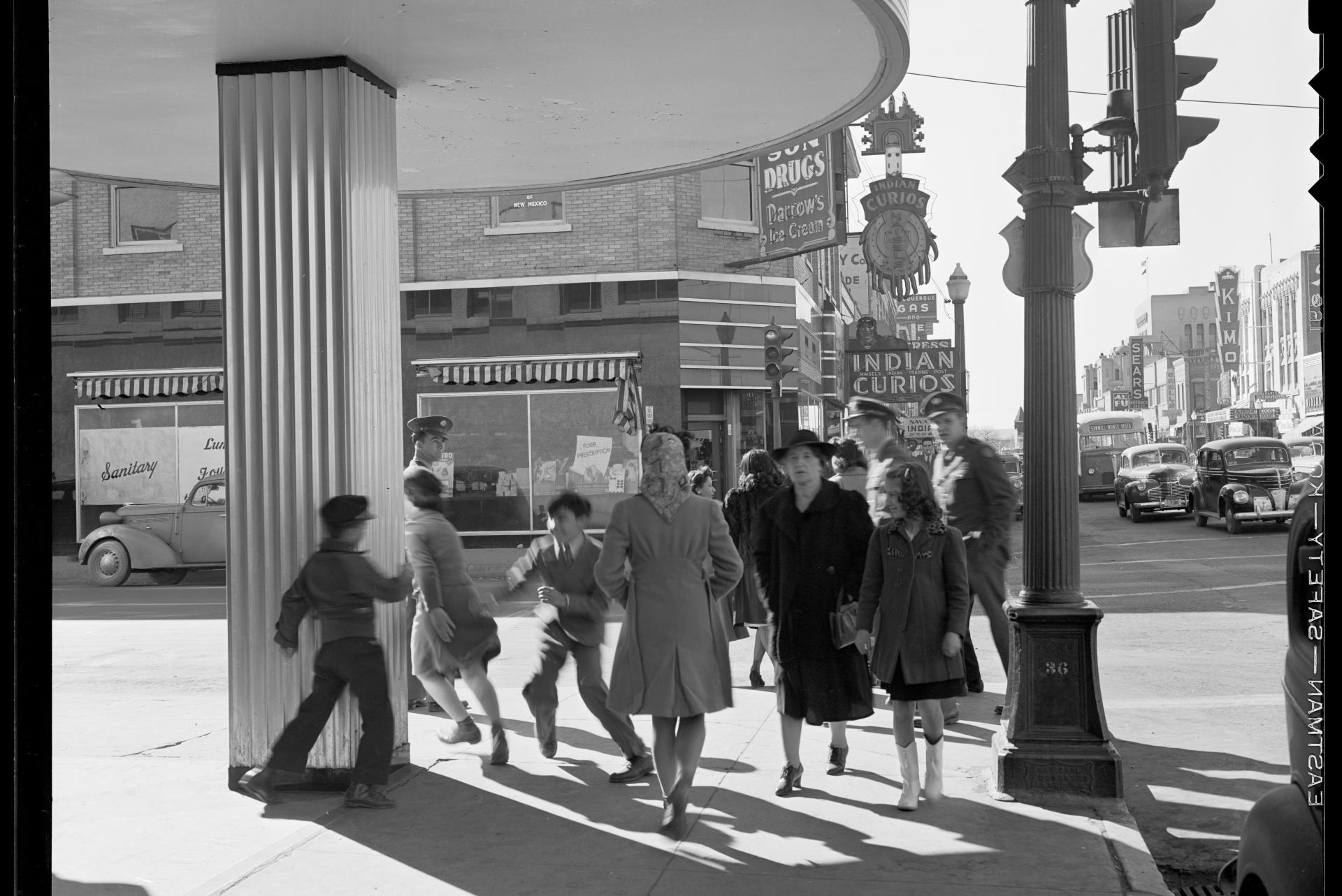 A bustling street corner scene shows people walking near a drug store with a visible sign. There are children playing and adults strolling past shops and parked cars. The storefronts display various signs, including one for Indian Curios. The setting appears to be from an earlier era, with vintage cars and fashion styles.