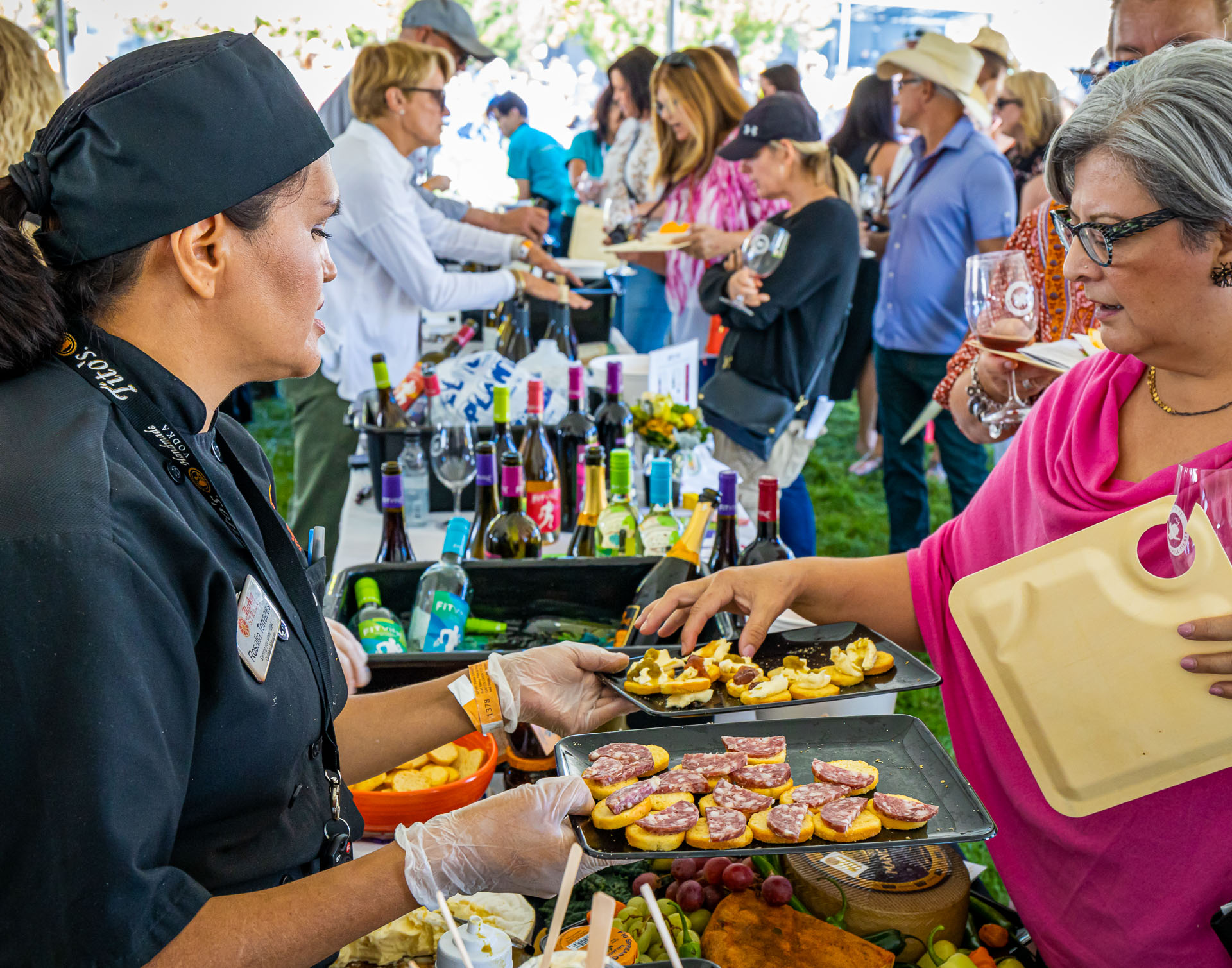 A bustling outdoor event with people gathered around tables featuring assorted foods and wine bottles. A person in a black chef's outfit serves appetizers to an attendee holding a wine glass. 