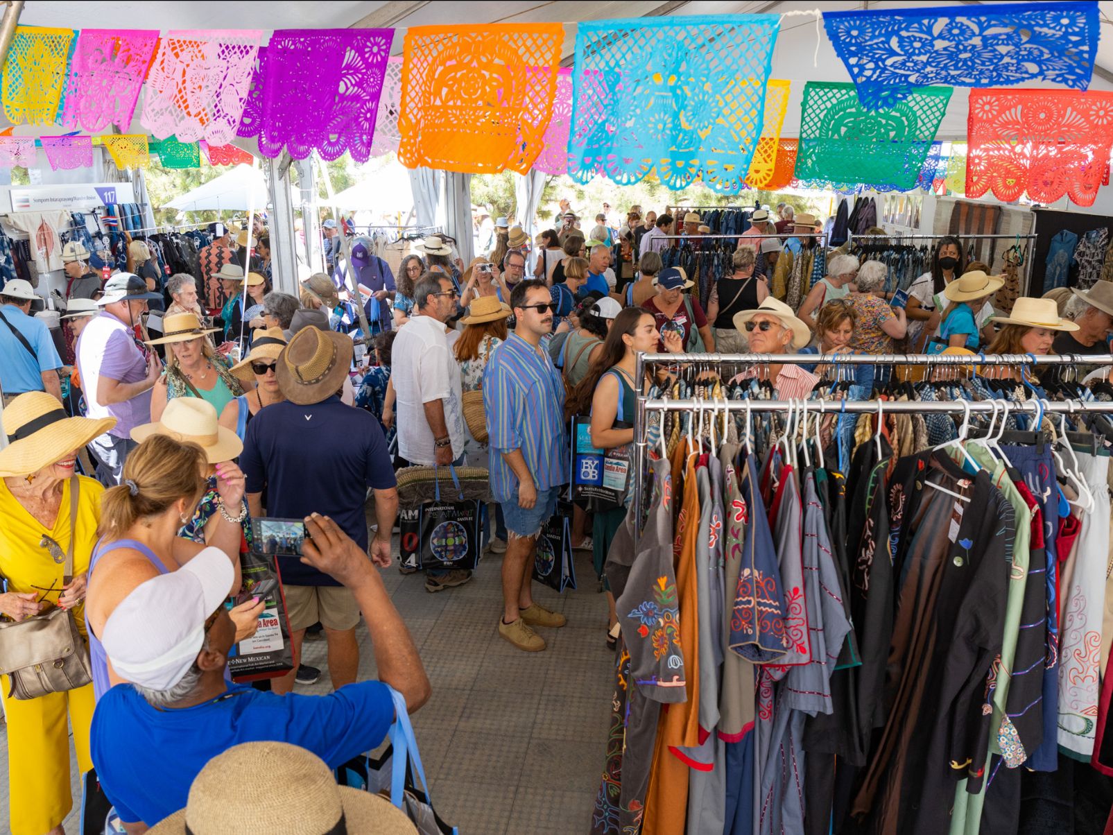 A crowd of people browse racks of clothing under an outdoor tent with colorful flags hanging from the ceiling