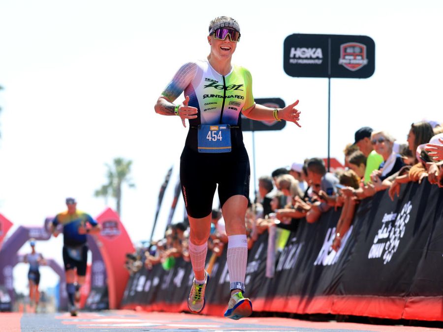 A person wearing athletic gear crosses a finish line in a race, smiling and spreading their arms. The backdrop includes banners with logos, such as HOKA, and a cheering crowd.