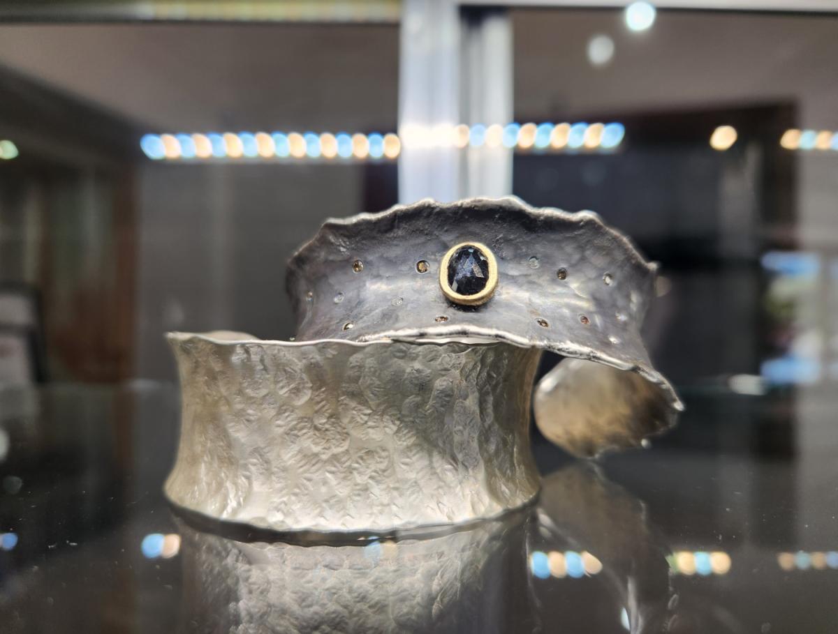 A textured silver bracelet with a wide band is on display. The bracelet features an oval-shaped black gemstone at its center. The background shows a softly lit display case.