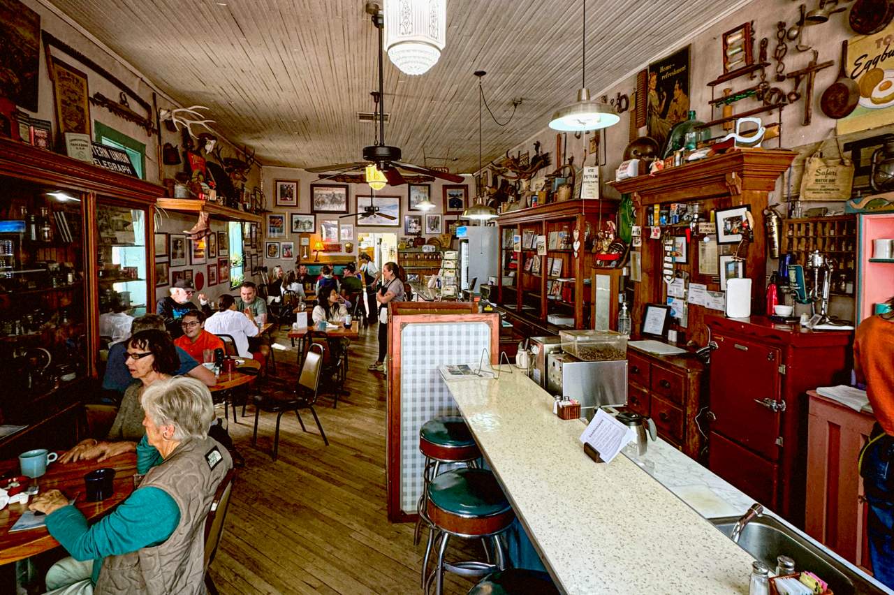This image shows a cozy, vintage-style diner with wooden floors and a well-worn bar. Patrons are seated at tables and the bar, enjoying meals and conversation. The walls are adorned with various memorabilia, photographs, and signs, creating a nostalgic ambiance.