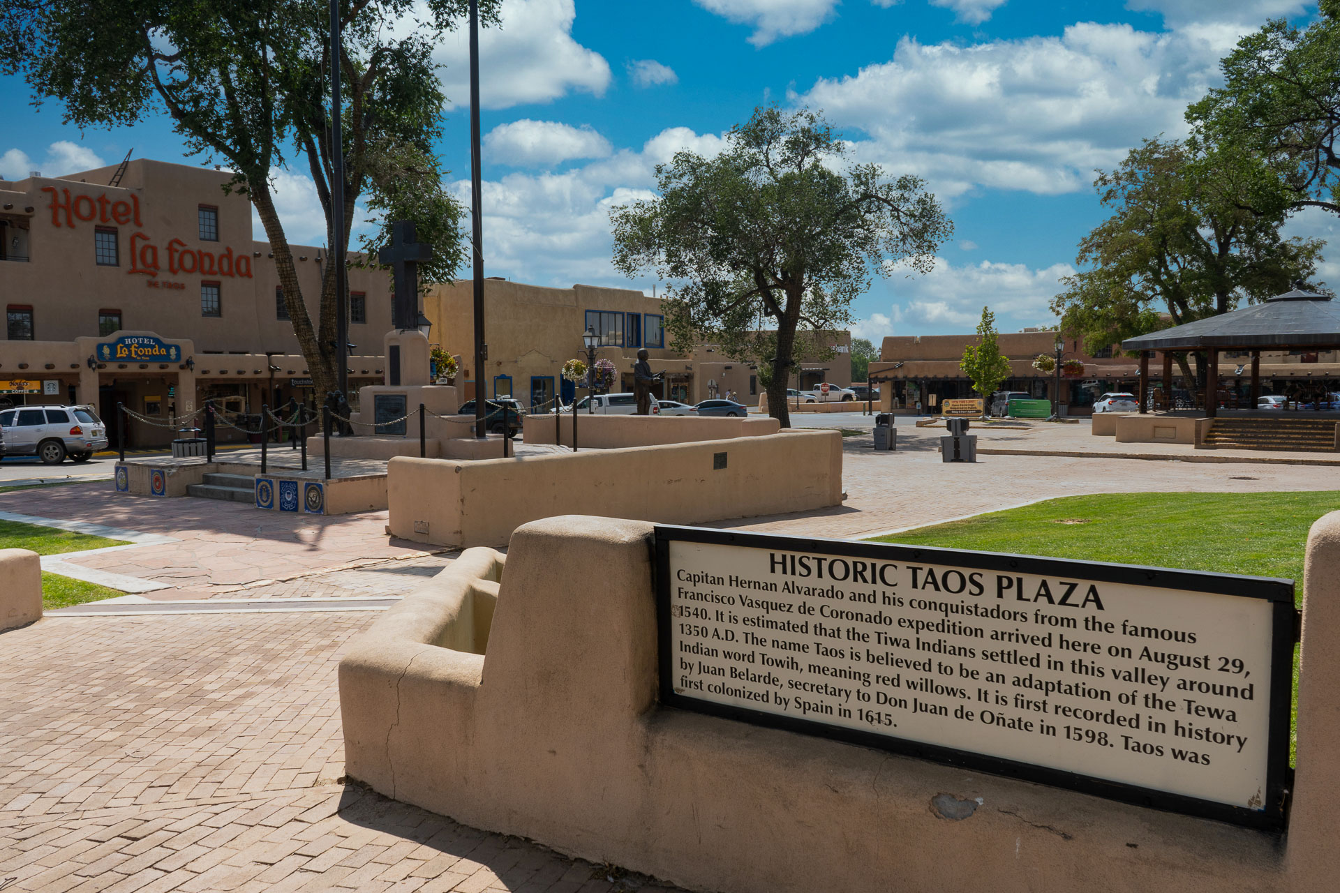 The Historic Taos Plaza on a sunny day, featuring a central open space bordered by pathways and greenery. The plaza is surrounded by traditional adobe-style buildings, including the Hotel La Fonda. A historical sign provides information about the plaza's origins and significance.