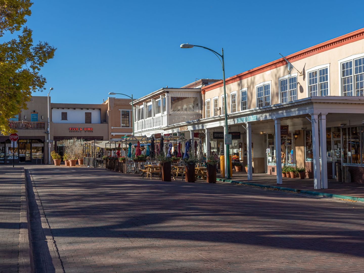 A sunny street scene in Santa Fe, New Mexico, features historic adobe buildings with wooden beams. A row of shops display colorful garments and various items outside.