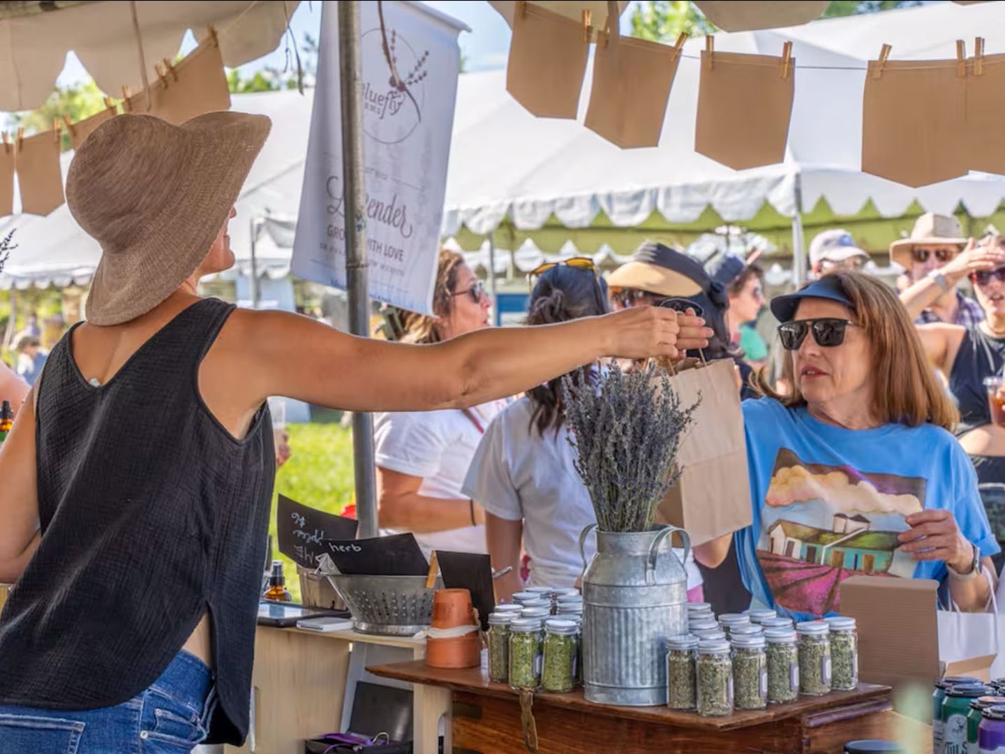 A person wearing a wide-brimmed hat and sleeveless top is handing a bag to another person with sunglasses and a colorful shirt at an outdoor market stall. The stall displays jars and a metal vase with lavender. There are multiple people and tents in the background.