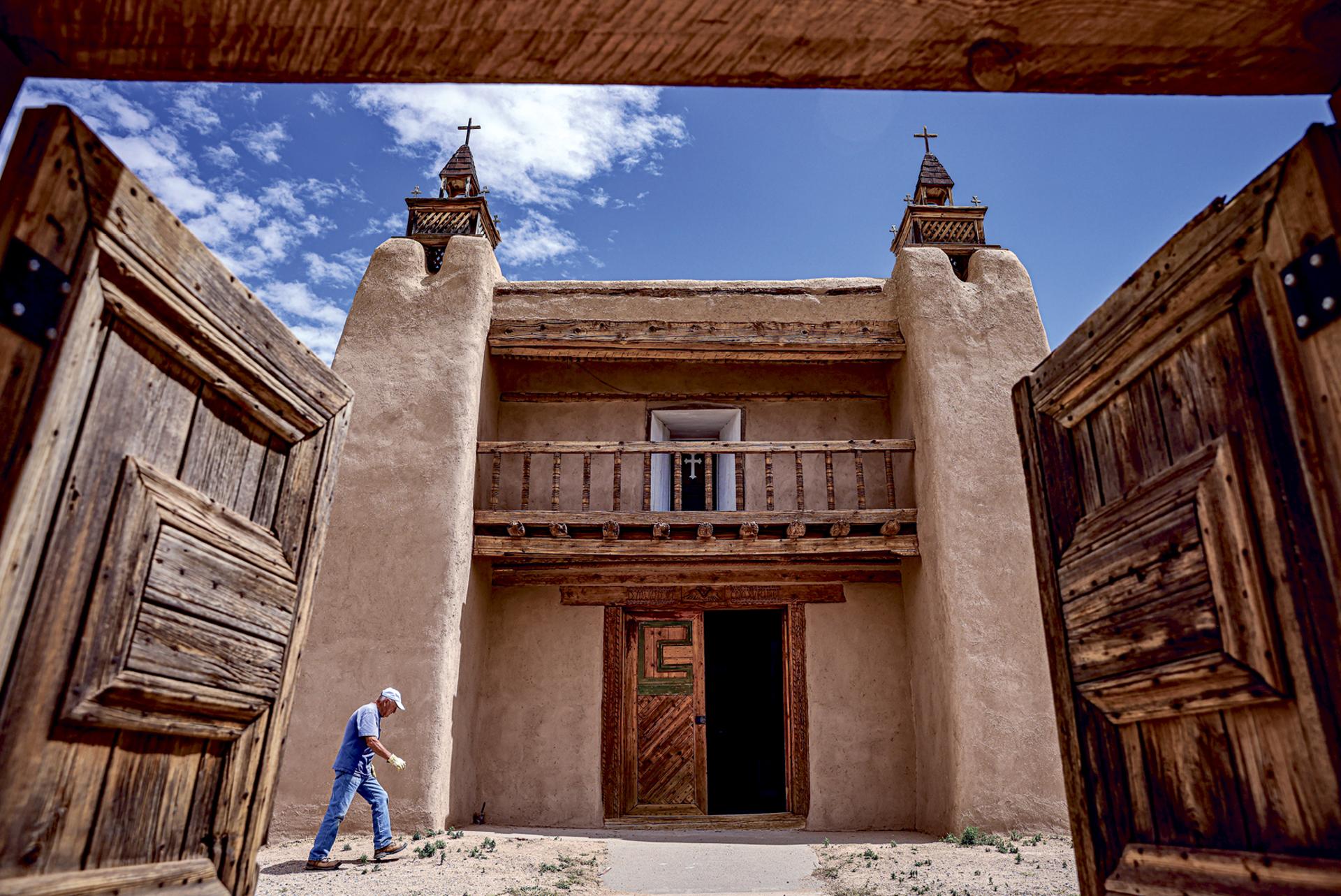 An old Spanish colonial church with wooden doors open in the foreground and a person walking nearby under a partly cloudy sky.
