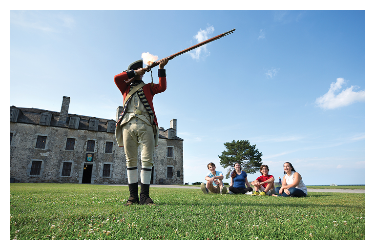 a man dressed in colonial attire shoots a rifle in the air as other people look on