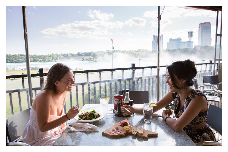 two women enjoying lunch on an outdoor porch with Niagara Falls in the background