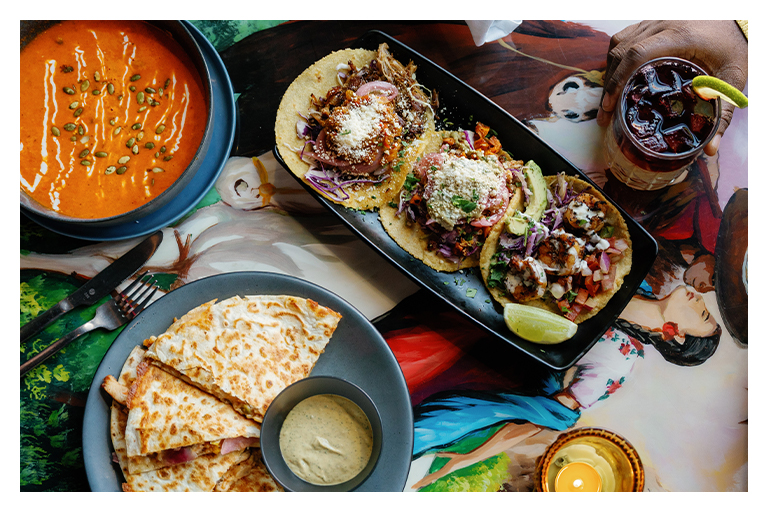 Plated tacos, quesadilla, and soup served on a colorful table, with a drink alongside.