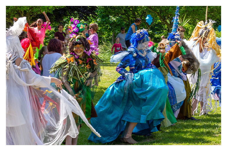 Colorful performers in flowing costumes dancing during a lively outdoor festival in a park.