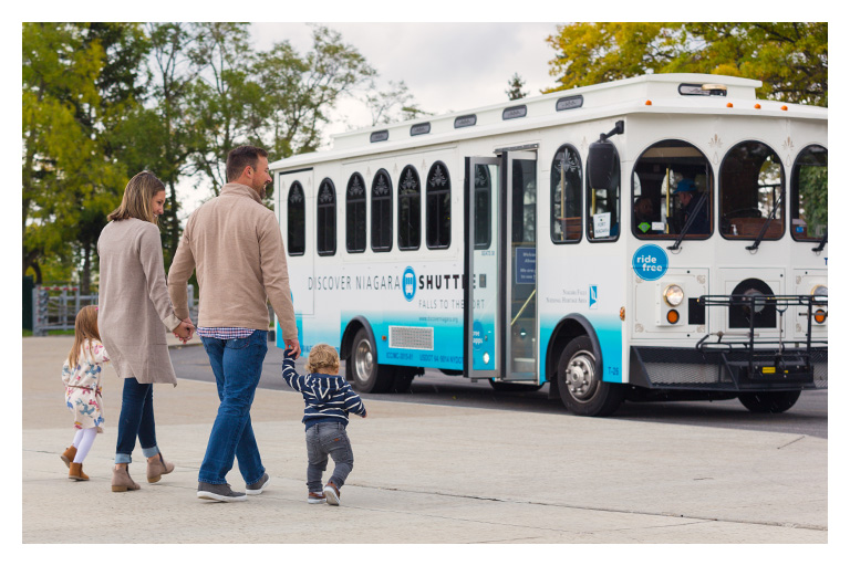 Family with young children walking toward a Discover Niagara Shuttle trolley on a tree-lined street.
