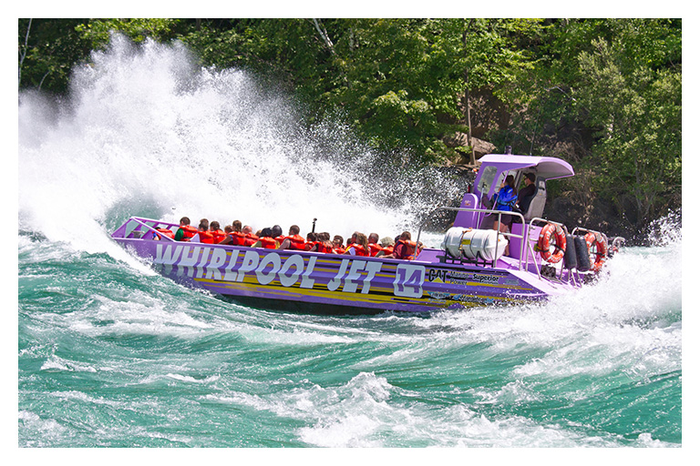 A Whirlpool Jet Boat Tours vessel speeds through the powerful Class V rapids of the Niagara River, sending waves crashing over excited passengers.