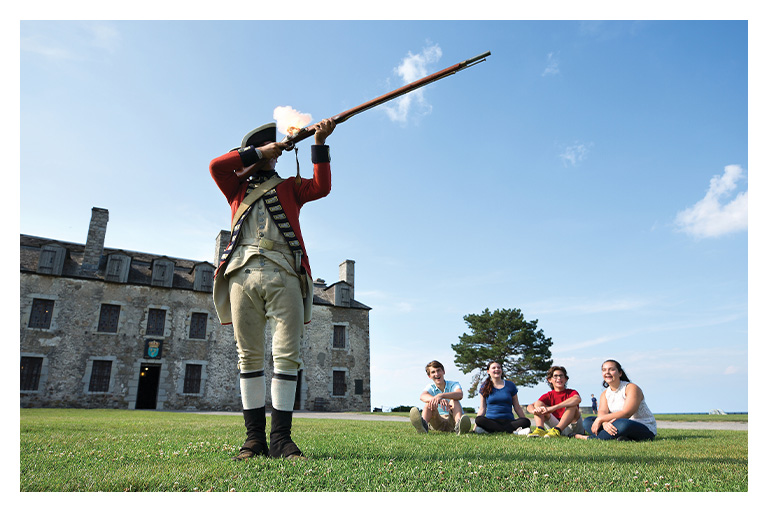 A historical reenactor fires a musket while visitors watch outside Old Fort Niagara on a bright summer day.