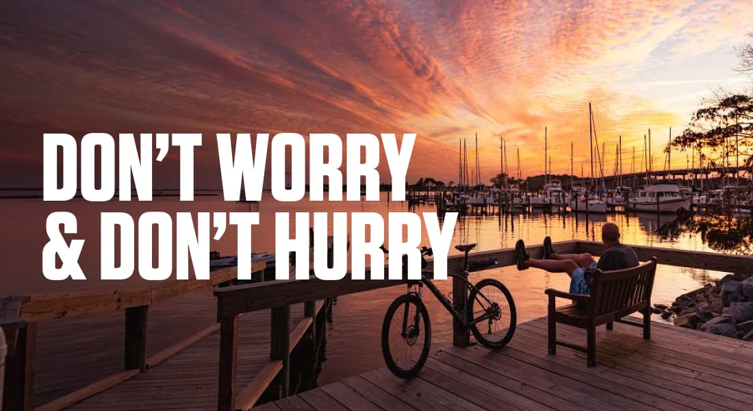 DON'T WORRY & DON'T HURRY - Man on bench on dock watching sunset over boats and water with his bike leaned up against railing