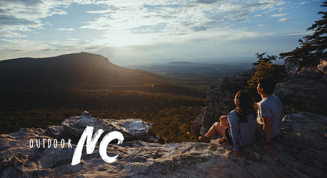 OUTDOOR NC - Young couple sitting on a hillside watching a sunrise over distant mountains