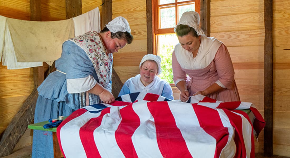 Three women dressed in historic outfits sewing a large American flag, recreating a scene from revolutionary times